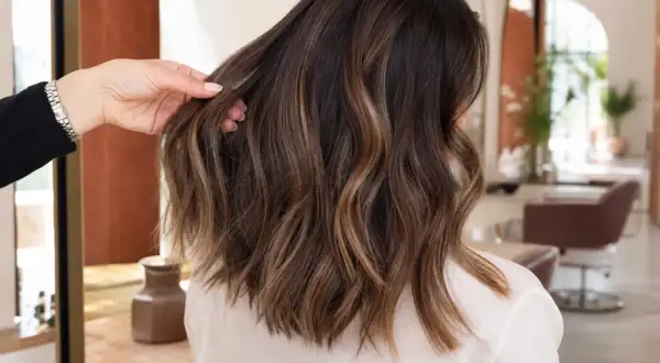 Une main ajuste une chevelure brune aux reflets subtils de balayage caramel, dans un salon de coiffure lumineux.