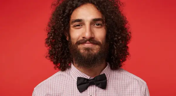 Homme souriant avec de longs cheveux bouclés, barbe et nœud papillon noir, sur fond rouge.
