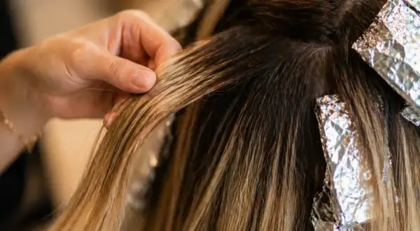 Main d'une coiffeuse travaillant sur des mèches de cheveux blonds, avec des feuilles d'aluminium. Le **foilyage vs balayage** est en cours.