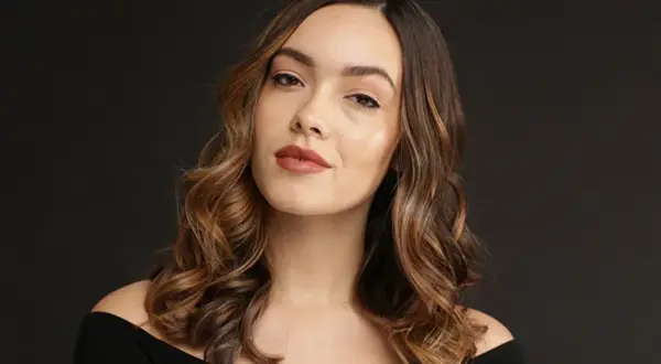 Une femme aux cheveux bruns ondulés, avec des reflets blonds, pose pour une photo. Elle a un maquillage naturel et un sourire subtil. Elle semble avoir réussi à faire un balayage maison.