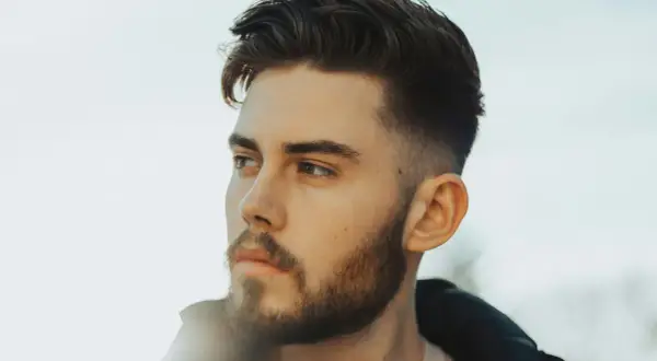 Jeune homme avec une barbe courte et une coupe de cheveux dégradée, style taper fade. Il regarde au loin, le visage éclairé par une lumière douce.