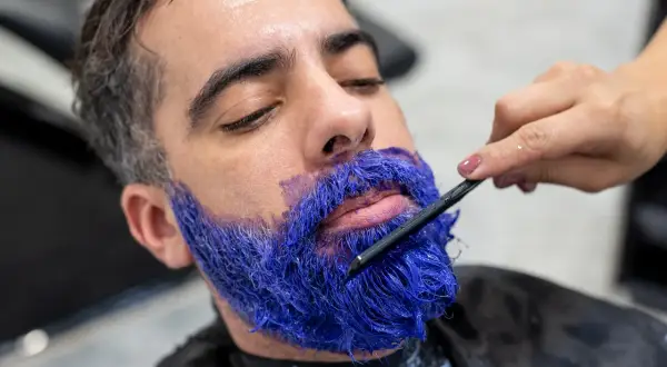 Un homme se fait teindre la barbe en bleu vif par une main tenant une brosse.