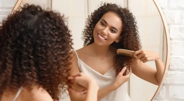 Une jeune femme souriante se regarde dans un miroir, peignant ses cheveux bouclés courts avec un peigne en bois.
