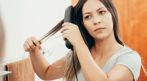 Une jeune femme réalise un lissage brésilien maison devant son miroir, utilisant un fer à lisser pour dompter ses cheveux.