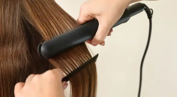 Une main utilise un fer à lisser pour lisser des cheveux bruns brillants, tandis qu'une autre main tient un peigne pour guider la mèche.