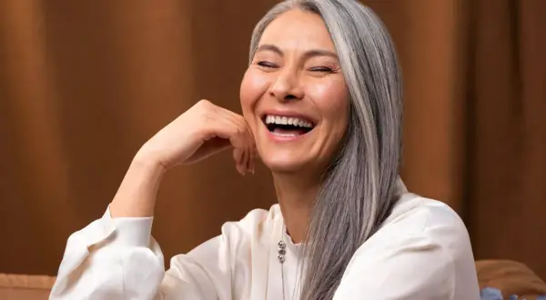 Une femme rit, sa main posée sur sa joue. Ses cheveux gris argenté ont des mèches de couleur subtiles, ajoutant une touche de modernité à son look.