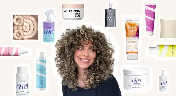 Une femme aux cheveux bouclés souriante, entourée de produits pour une routine cheveux bouclés : sprays, masques, gels et conditionneurs.