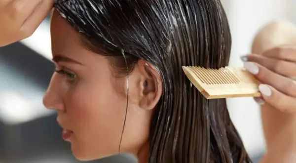Une femme applique un masque huile cheveux avec un peigne en bois. Ses cheveux sont longs et bruns, et le produit est visible sur les longueurs.