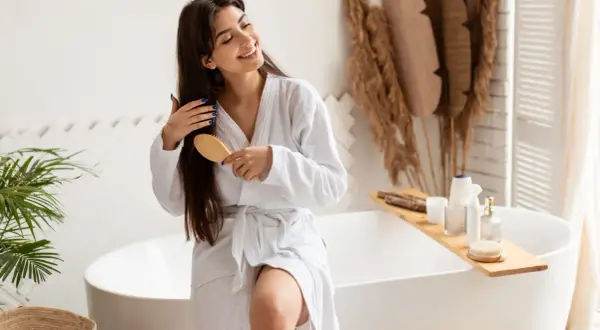 Une femme souriante en peignoir blanc se brosse les cheveux longs et bruns, un geste doux pour nourrir ses cheveux.