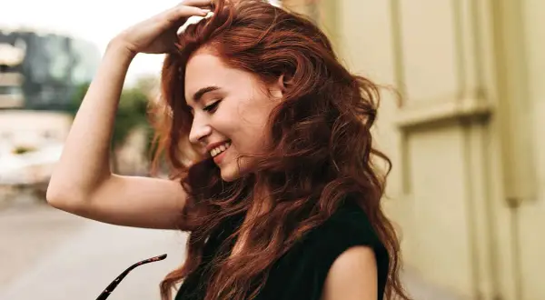 Une jeune femme aux cheveux roux, souriante, joue avec sa chevelure. Le **dosage coloration oxydant 30** a révélé cette belle teinte.