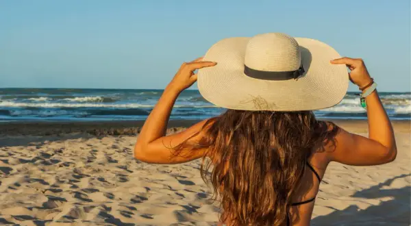 Une femme à la plage, coiffée d'un grand chapeau, montre comment protéger ses cheveux du soleil.