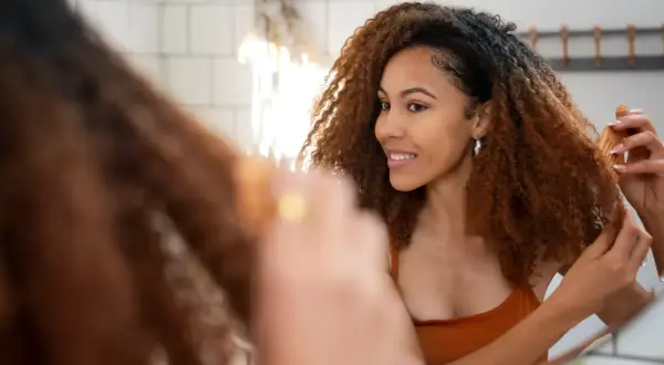 Une femme aux cheveux bouclés foncés se regarde dans le miroir, une brosse à la main. Elle rêve d'éclaircir cheveux noir sans décoloration.