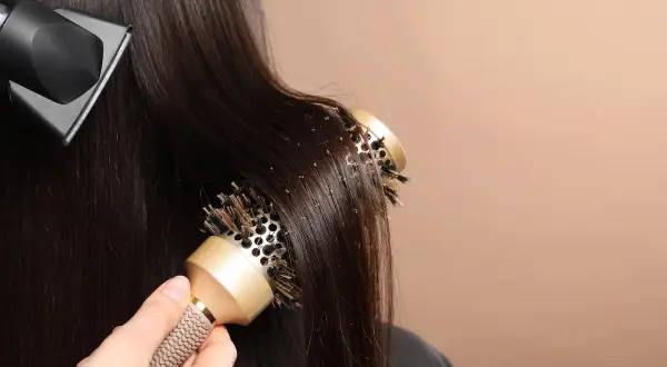 Une main utilise une brosse ronde pour coiffer des cheveux bruns, tandis qu'un sèche-cheveux souffle de l'air. Le brushing boucle est en cours.