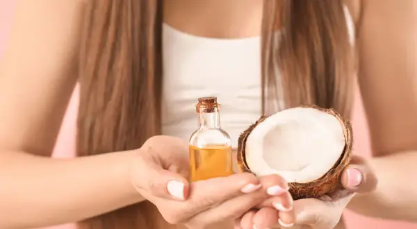 Une femme tient une petite bouteille d'huile dorée et une moitié de noix de coco. L'huile de coco cheveux est un secret de beauté naturel.