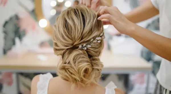 Une mariée se fait coiffer un chignon bas flou, orné d'une barrette scintillante. Les mains d'une coiffeuse ajustent les mèches.