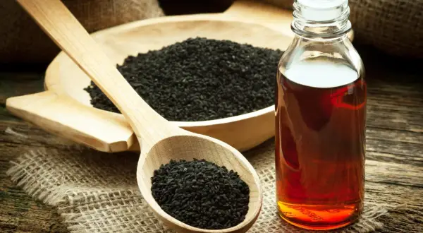 Une cuillère en bois remplie de graines de nigelle noire, à côté d'un bol de graines et d'une bouteille d'huile ambrée, parfaite pour l'huile de nigelle cheveux.