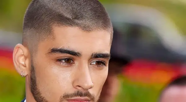Un jeune homme au regard intense arbore une coupe buzz, ses cheveux courts et nets accentuent son style moderne.