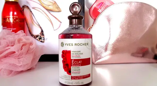 Bouteille de vinaigre de framboise pour cheveux Yves Rocher, avec des chaussures stylisées et une trousse de toilette rose en arrière-plan.