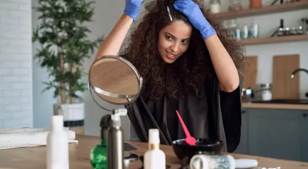 Une femme souriante fait sa coloration cheveux maison, appliquant le produit avec des gants bleus. Un miroir et des accessoires de coiffure sont sur la table.