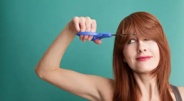 Une femme aux cheveux roux tente de comment couper une frange avec des ciseaux bleus.