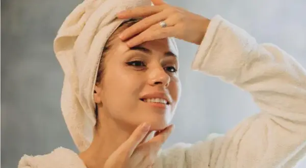 Une femme avec une serviette sur la tête et un peignoir, se touchant le visage. Une astuce pour savoir comment faire une serviette chaude pour les cheveux.