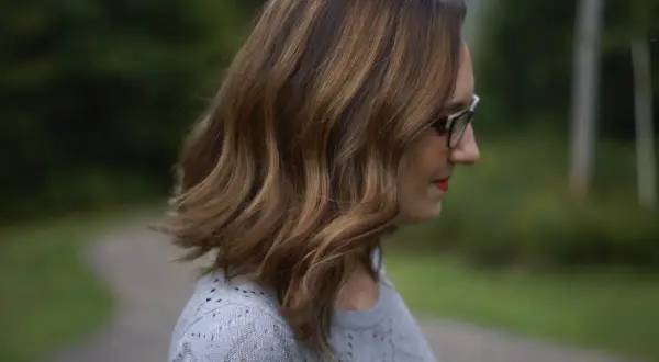 Une femme aux mèches brunes sur cheveux blancs, portant des lunettes, regarde au loin dans un décor verdoyant.