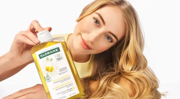 Une femme aux cheveux blonds tient un flacon de shampoing Klorane à la camomille, un produit pour cheveux éclaircissant.