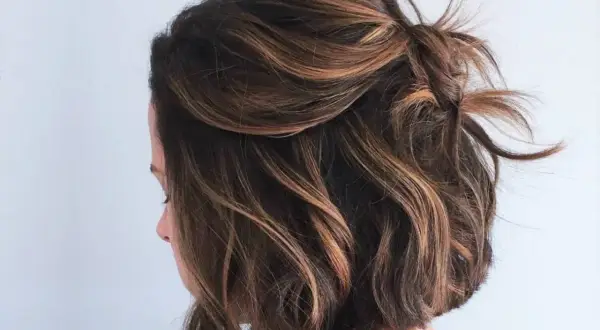 Coupe courte ondulée avec des reflets blonds subtils, le dessus est relevé en un chignon flou.