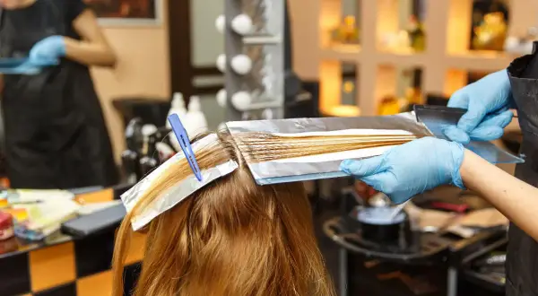 Coiffure : une main gantée applique une décoloration sur des mèches fines, une technique pour comment faire ses racines quand on a des mèches.