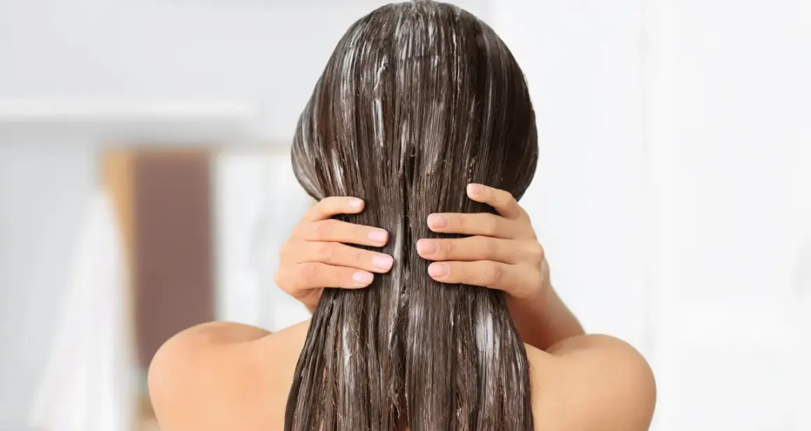 Application d'un masque pour sceller l'hydratation des cheveux. Les mains massent le produit sur les longueurs brunes et humides.