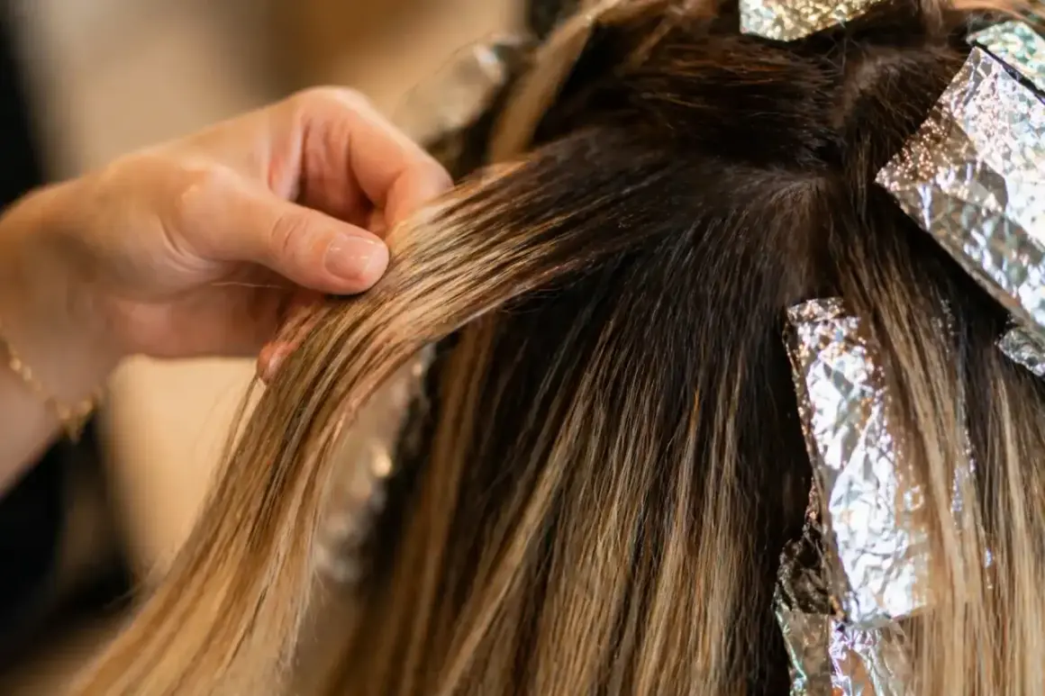 Main d'une coiffeuse travaillant sur des mèches de cheveux blonds, avec des feuilles d'aluminium. Le **foilyage vs balayage** est en cours.