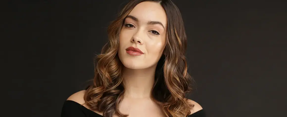 Une femme aux cheveux bruns ondulés, avec des reflets blonds, pose pour une photo. Elle a un maquillage naturel et un sourire subtil. Elle semble avoir réussi à faire un balayage maison.