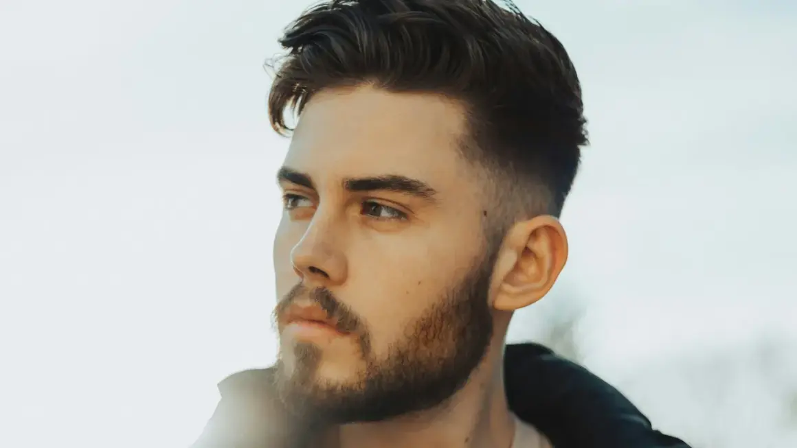 Jeune homme avec une barbe courte et une coupe de cheveux dégradée, style taper fade. Il regarde au loin, le visage éclairé par une lumière douce.