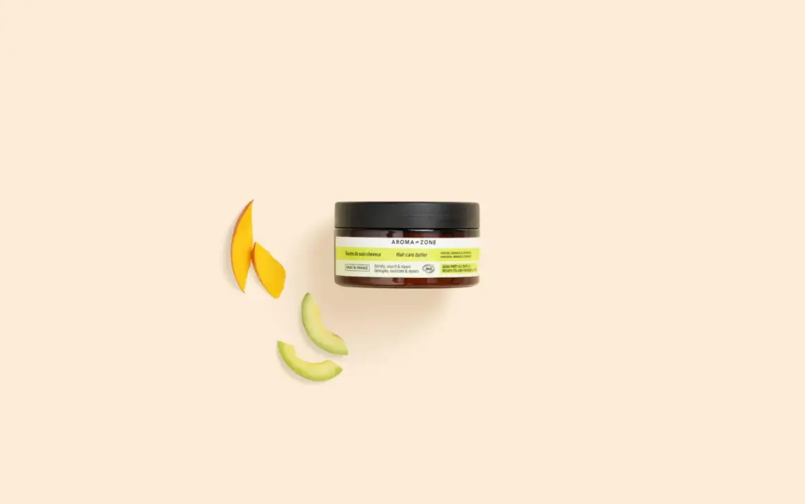 Un pot de beurre de soin cheveux Aroma-Zone, avec des tranches de mangue et d'avocat à côté.