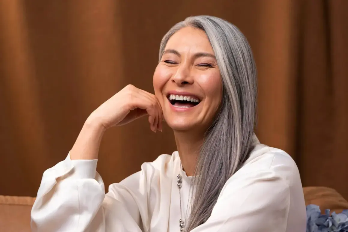 Une femme rit, sa main posée sur sa joue. Ses cheveux gris argenté ont des mèches de couleur subtiles, ajoutant une touche de modernité à son look.