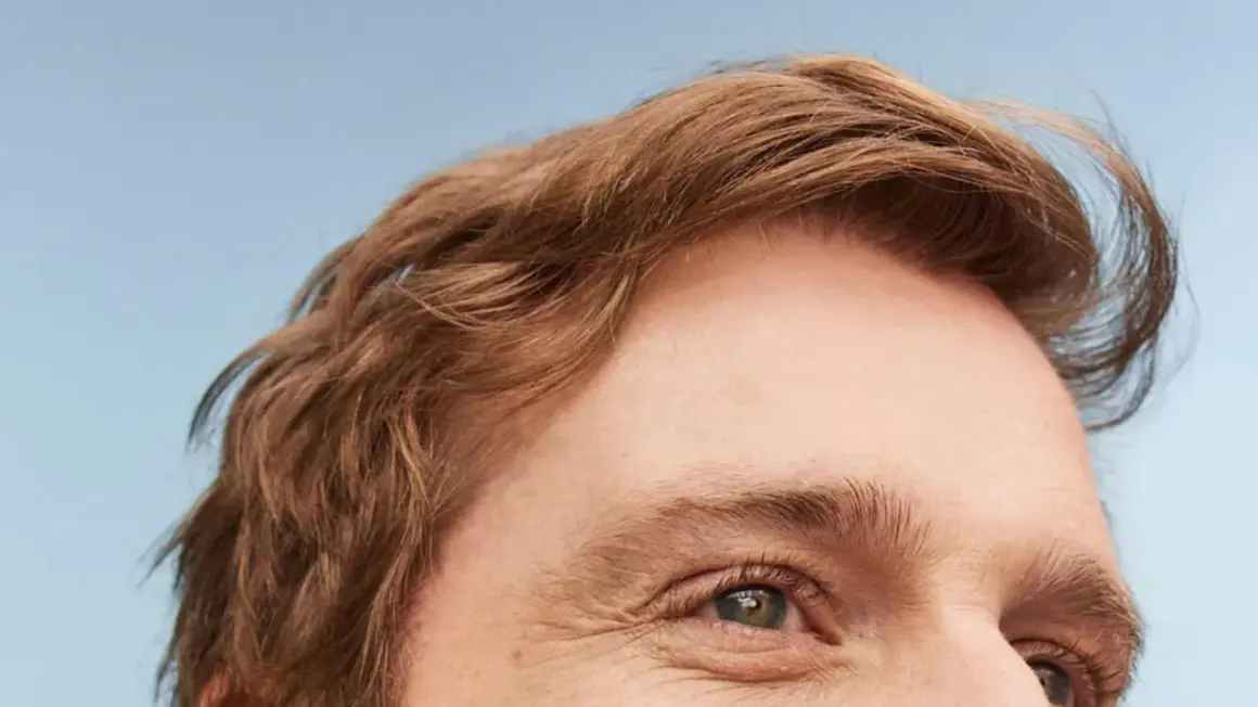 Un homme au regard clair, ses cheveux châtains légèrement ondulés, un brushing naturel.