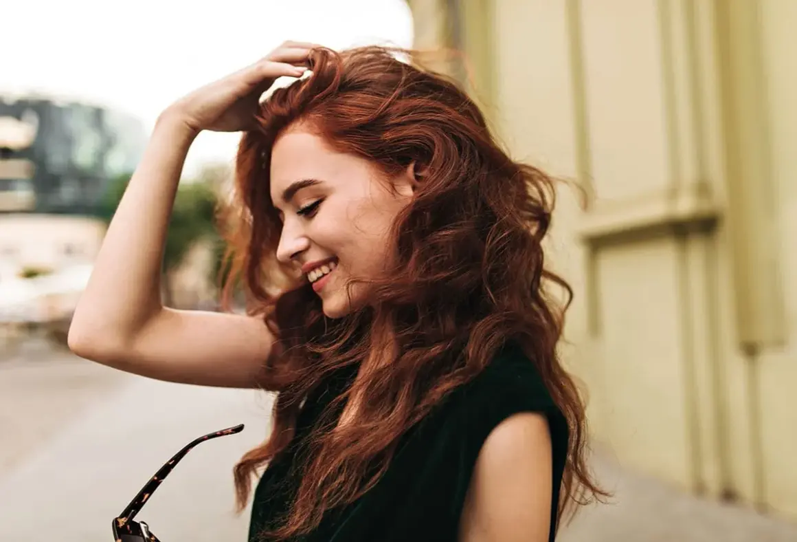 Une jeune femme aux cheveux roux, souriante, joue avec sa chevelure. Le **dosage coloration oxydant 30** a révélé cette belle teinte.