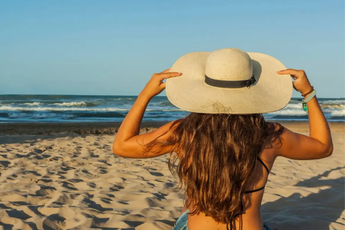 Une femme à la plage, coiffée d'un grand chapeau, montre comment protéger ses cheveux du soleil.