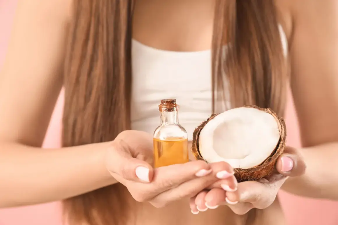 Une femme tient une petite bouteille d'huile dorée et une moitié de noix de coco. L'huile de coco cheveux est un secret de beauté naturel.