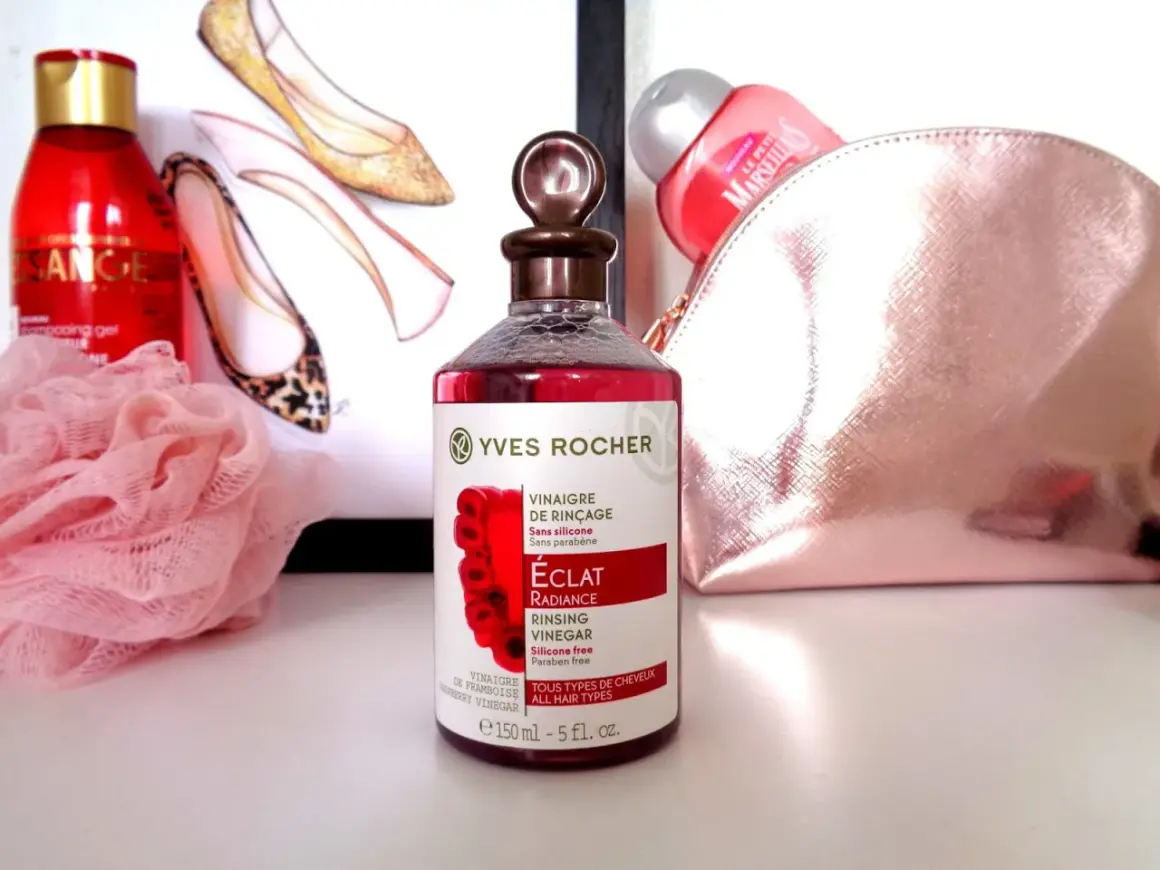 Bouteille de vinaigre de framboise pour cheveux Yves Rocher, avec des chaussures stylisées et une trousse de toilette rose en arrière-plan.