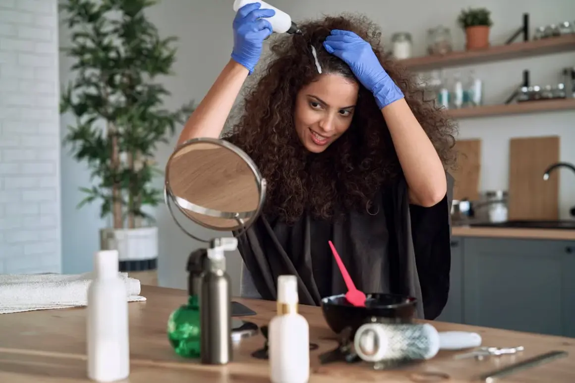 Une femme souriante fait sa coloration cheveux maison, appliquant le produit avec des gants bleus. Un miroir et des accessoires de coiffure sont sur la table.