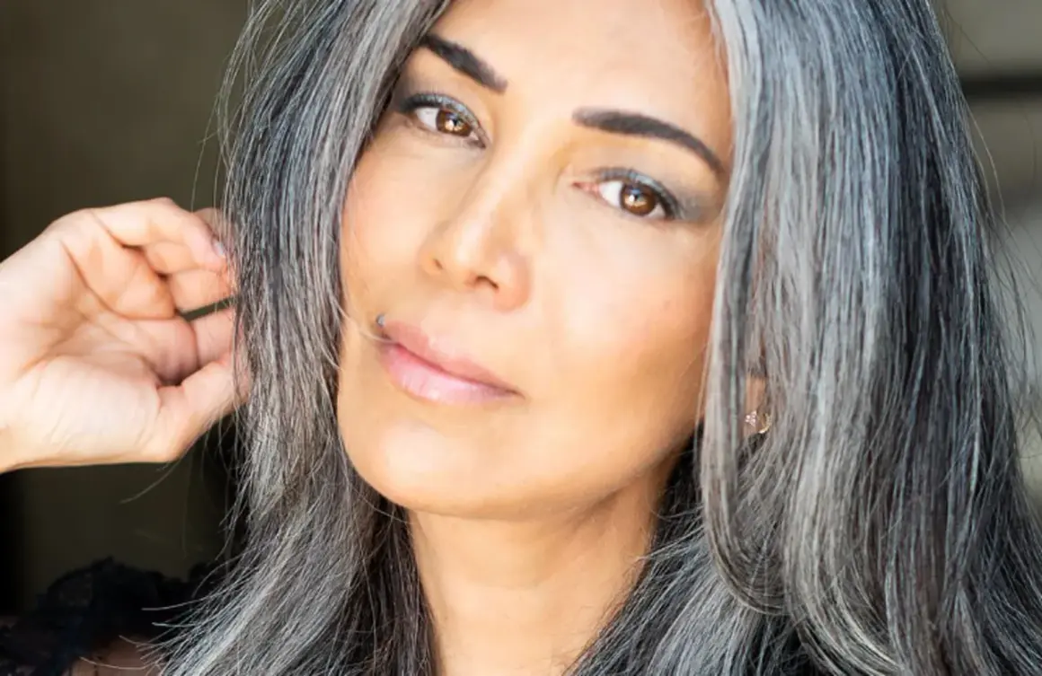 Une femme avec des cheveux gris mi-courts, le regard intense, touche doucement sa chevelure.