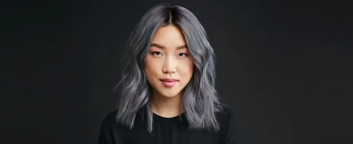 Une jeune femme au teint clair arbore une chevelure mi-longue, ondulée et d'un magnifique gris argenté. Elle porte un haut noir.