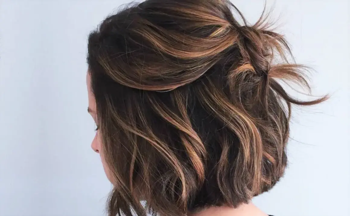 Coupe courte ondulée avec des reflets blonds subtils, le dessus est relevé en un chignon flou.