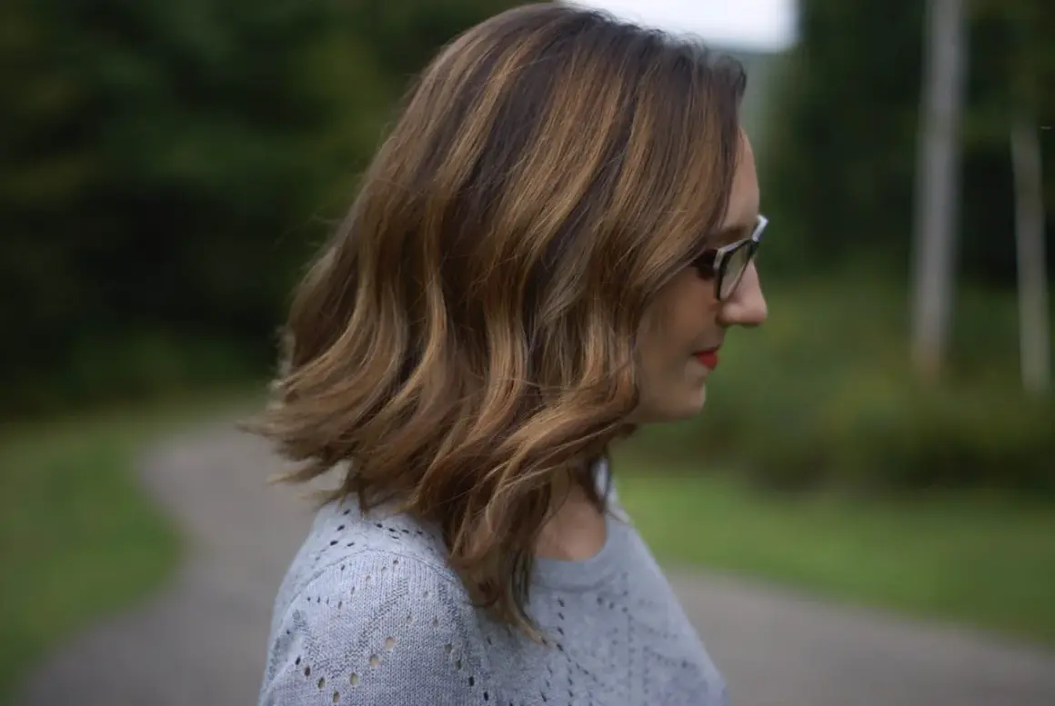 Chevelure châtain avec reflets blonds, une femme porte des lunettes. Une idée pour comment cacher ses cheveux blancs quand on est blonde.