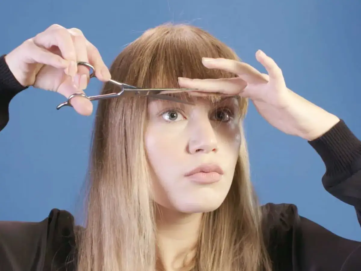 Une femme se coupe la frange avec des ciseaux. Elle tient ses cheveux avec sa main gauche et coupe avec sa main droite.