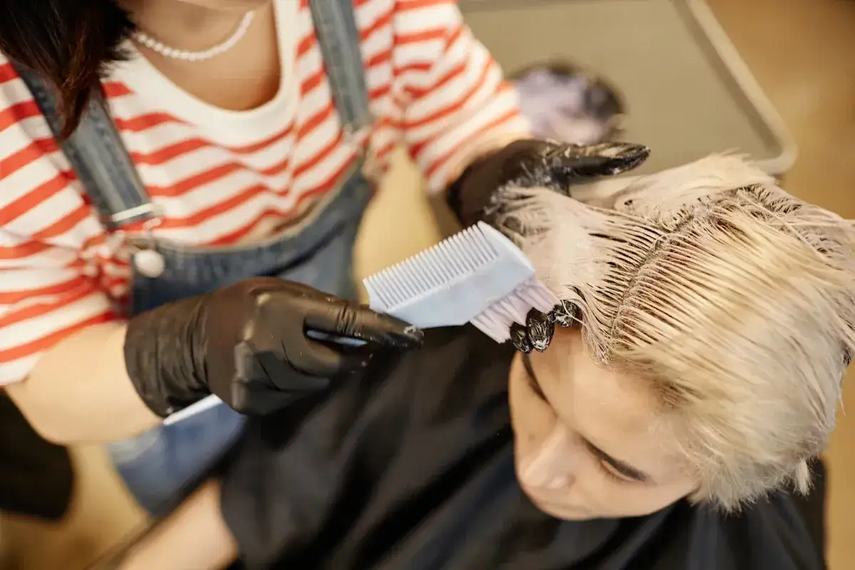 Application d'une patine sur cheveux colorés blonds par une coiffeuse portant des gants noirs.