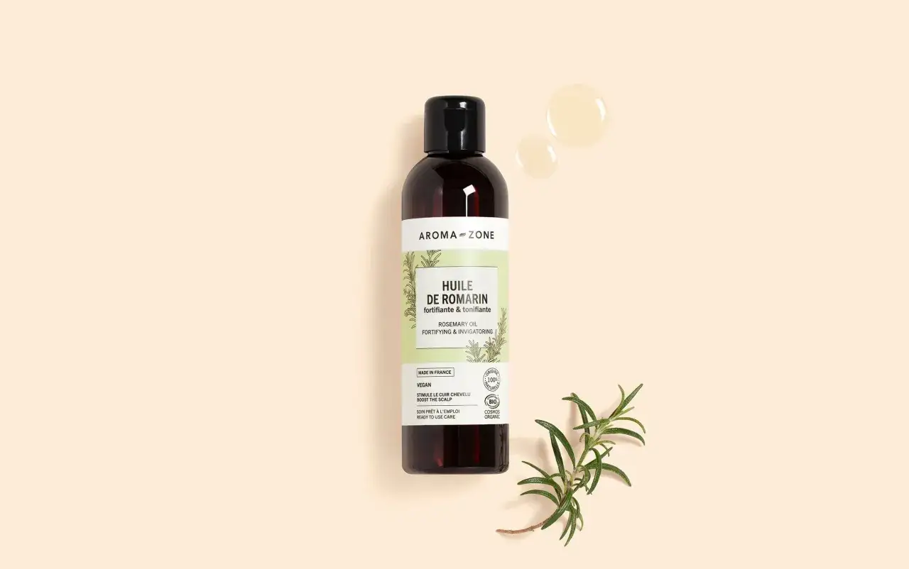 Huile de romarin Aroma-Zone pour stimuler la pousse des cheveux. Une goutte d'huile et une branche de romarin sur fond beige.
