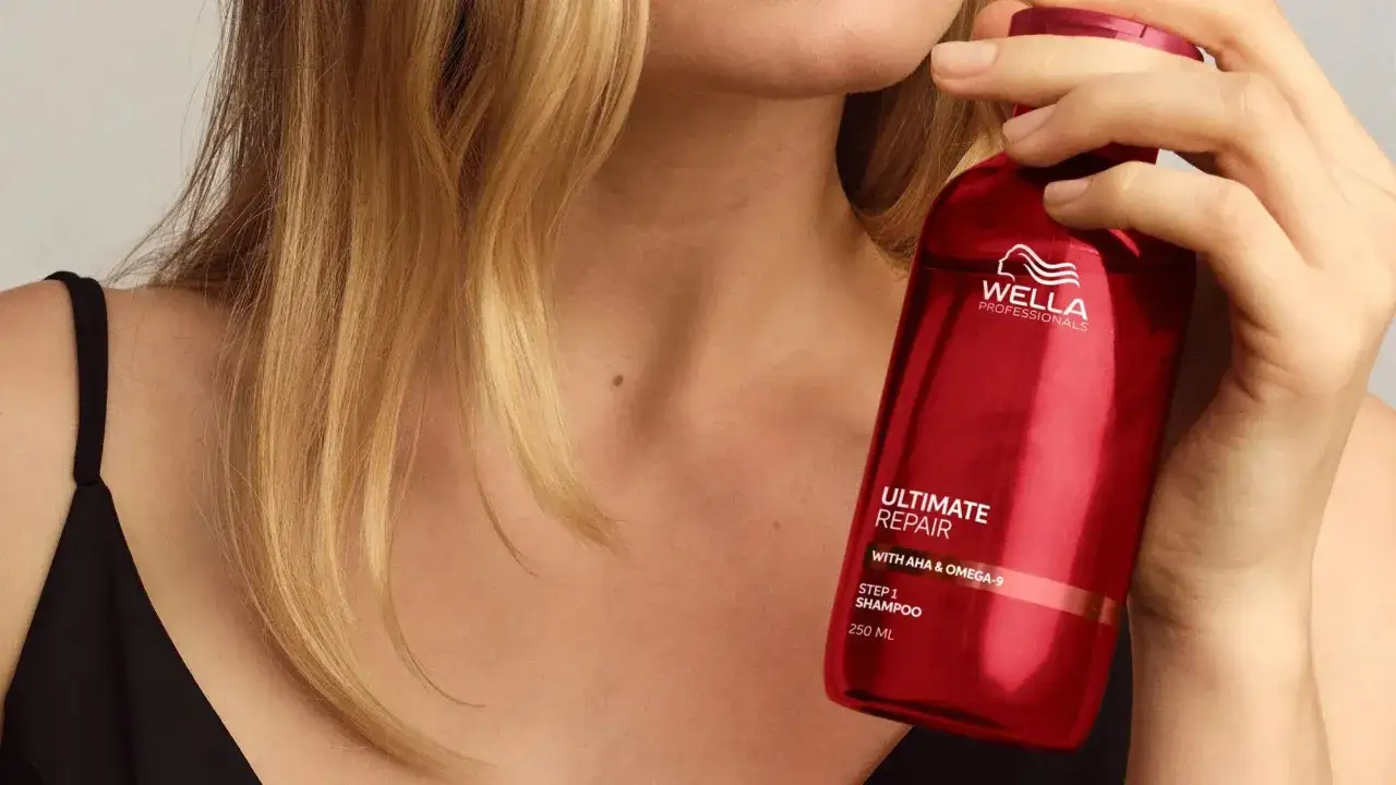 Une femme aux cheveux blonds tient une bouteille rouge de shampoing Wella Ultimate Repair, le premier shampoing après décoloration pour réparer les cheveux abîmés.