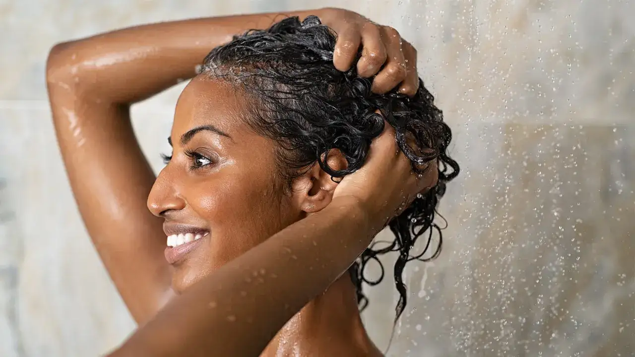Une femme souriante se lave les cheveux sous la douche. Elle a les cheveux fonc&eacute;s et ondul&eacute;s. Elle semble appr&eacute;cier ce moment, peut-&ecirc;tre une fois toutes les deux semaines.
